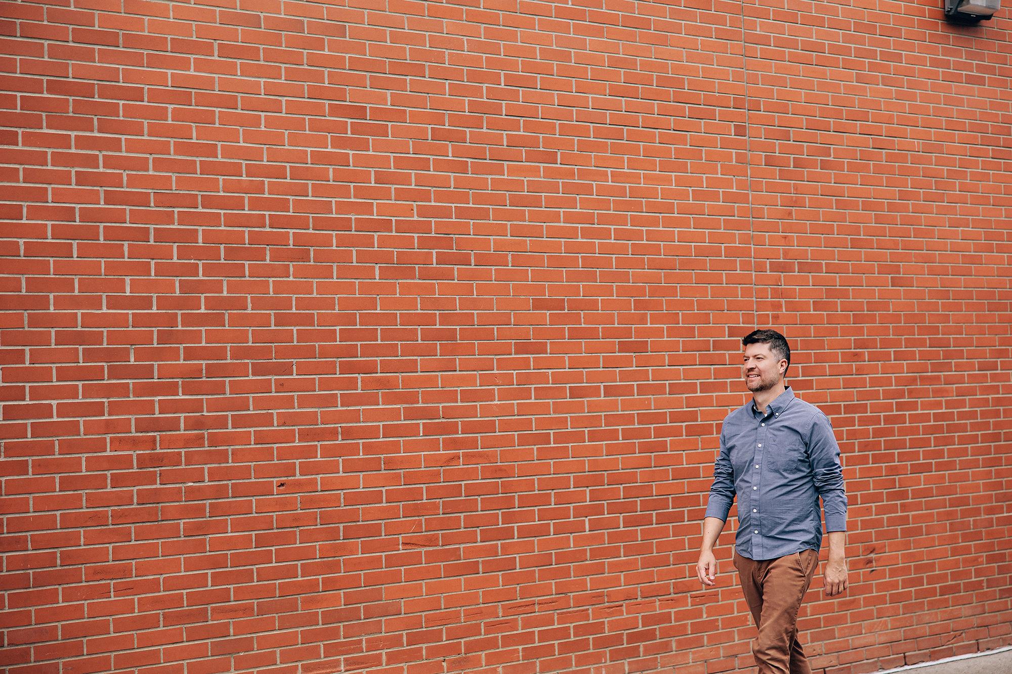 A smiling man in a blue button-down shirt walks past a textured red brick wall, showcasing a casual personal branding style by a Calgary photographer.