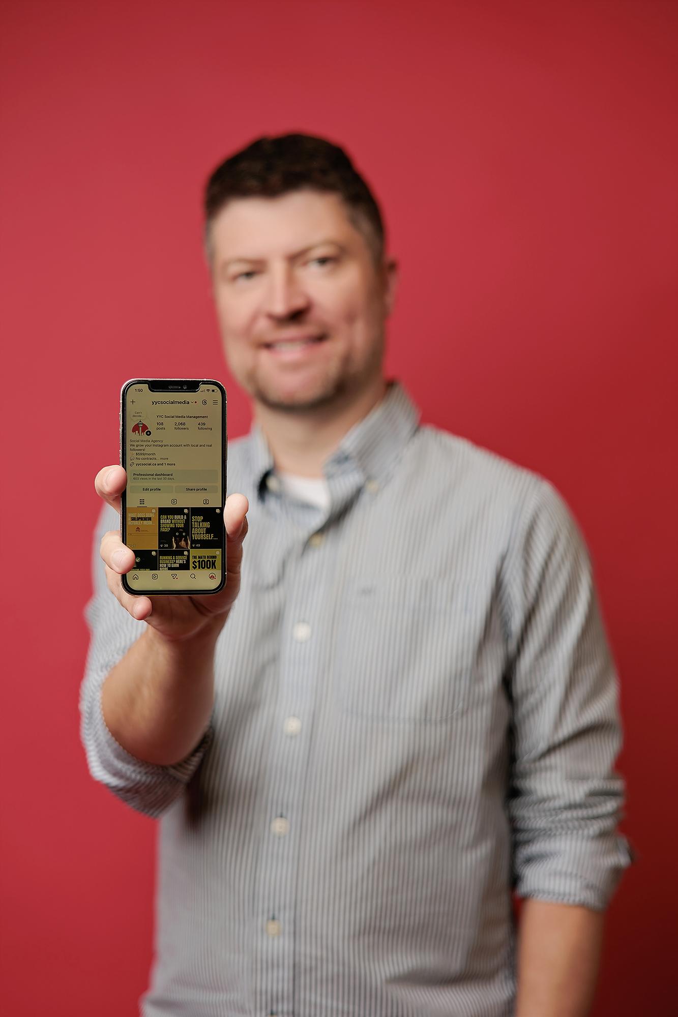 A smiling man in a striped shirt holds a smartphone displaying a social media profile, captured at our Calgary photography studio for personal branding.