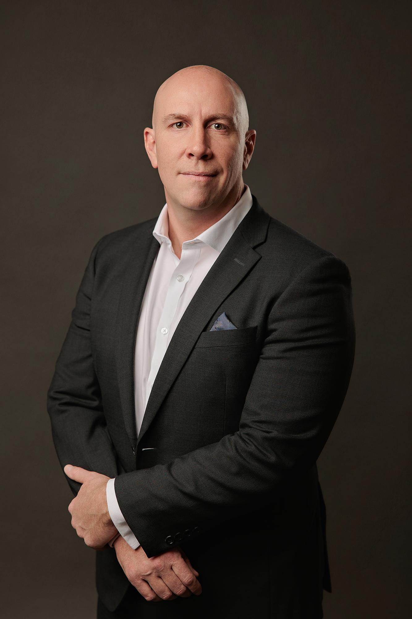 Professional headshots Calgary studio: Authoritative bald male executive in a dark charcoal suit and white dress shirt with a blue pocket square, standing with hands clasped and a composed expression against a warm dark charcoal background