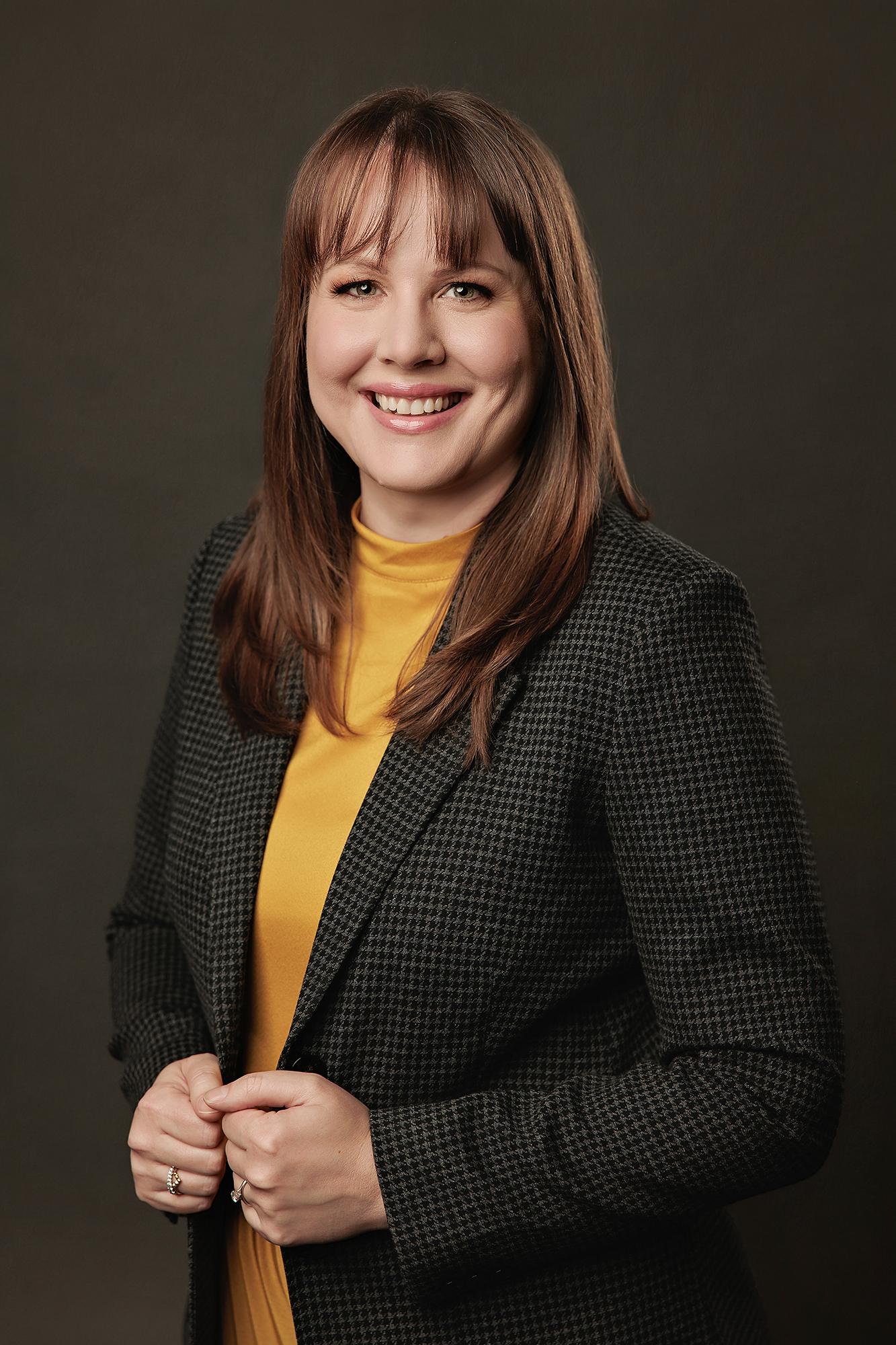 Professional headshots Calgary studio: Warm and approachable woman with auburn hair and wispy bangs in a dark houndstooth blazer over a mustard yellow turtleneck, hands clasped with a bright smile against a warm dark charcoal background