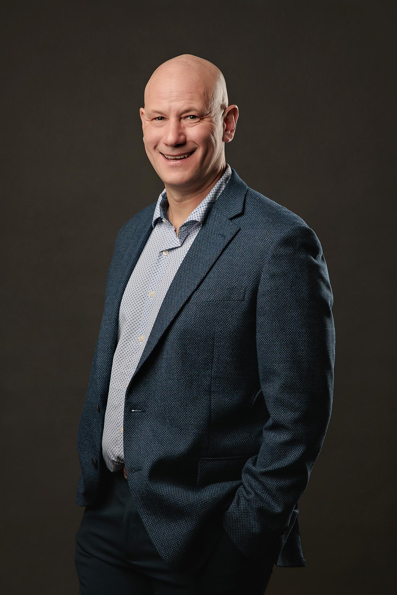 Professional headshots Calgary studio: Energetic bald male professional in a teal textured blazer and light blue dot-print shirt with hands in pockets and a broad smile in a relaxed three-quarter pose against a warm dark charcoal background
