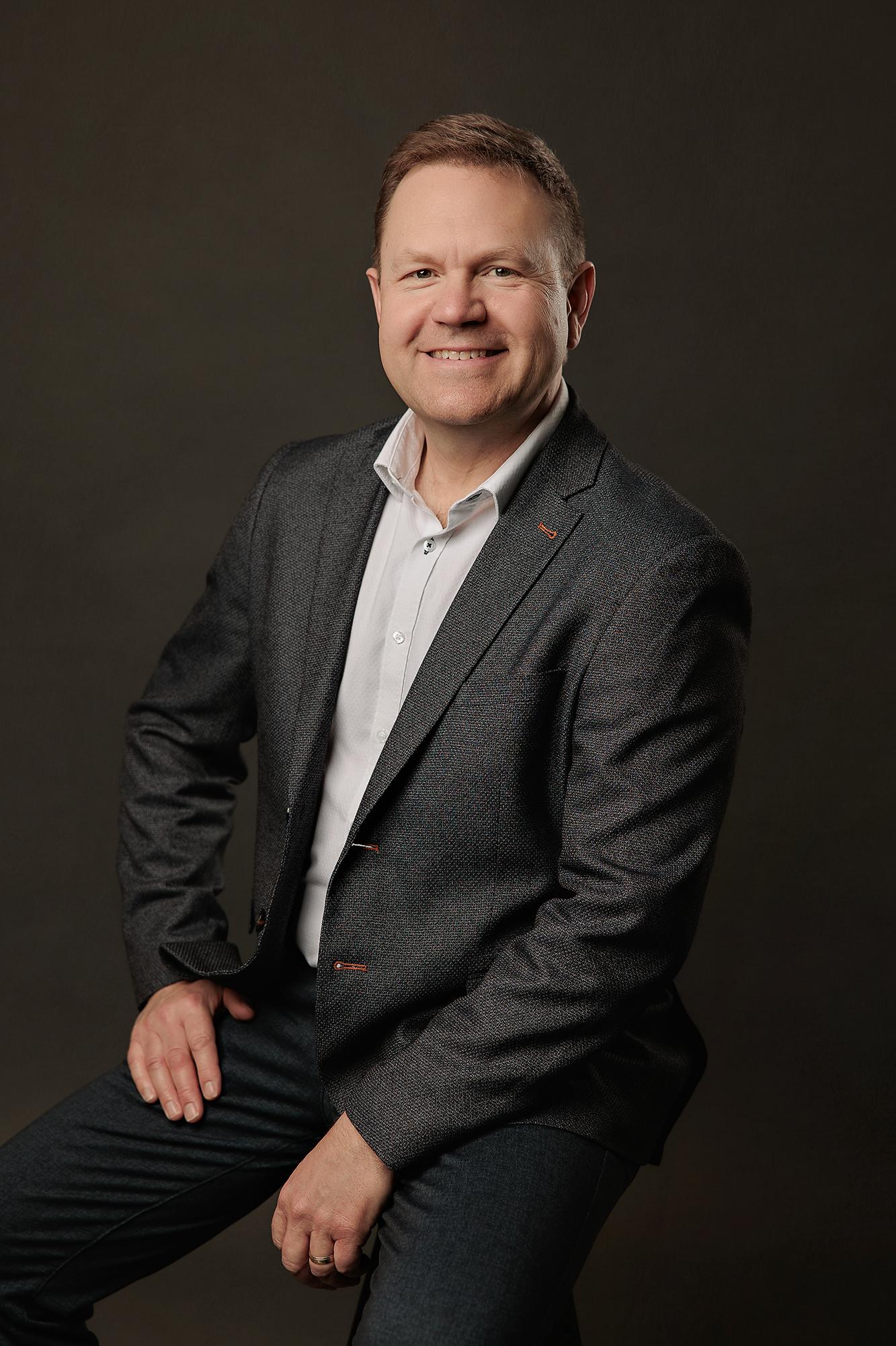 Professional headshots Calgary studio: Charismatic male professional with short salt-and-pepper hair in a dark grey fleck blazer with orange accent stitching and a white dress shirt, seated and leaning forward with a broad smile against a warm dark charcoal background