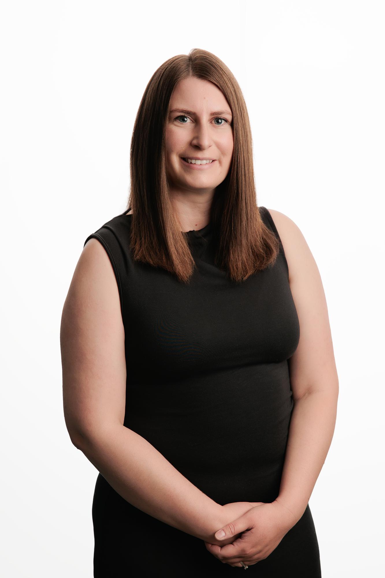 Team headshots Calgary studio: Approachable professional woman with straight brown hair in a black sleeveless top with hands clasped against a white studio backdrop