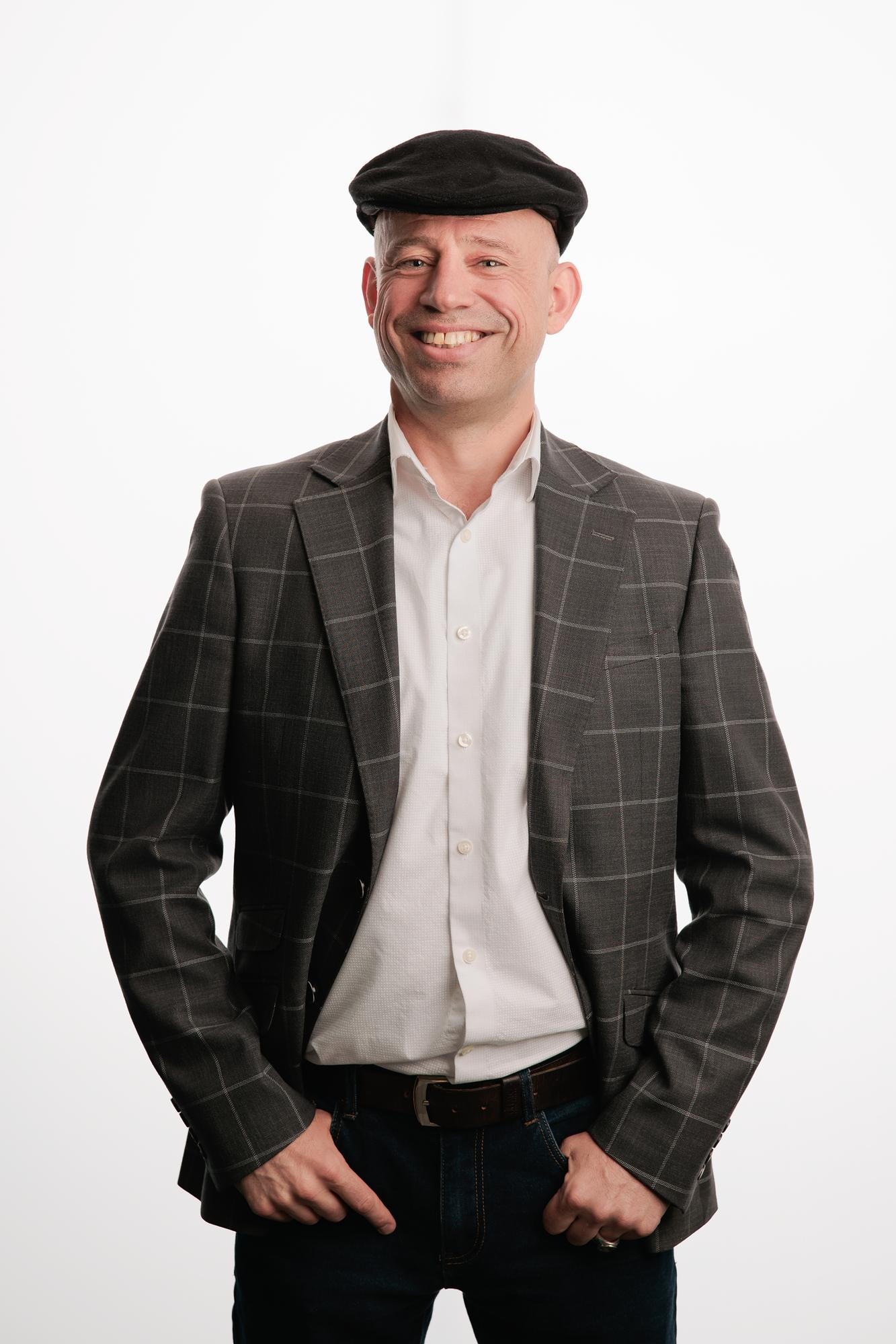 Team headshots Calgary studio: Full-length portrait of a professional man in a grey plaid windowpane blazer, white shirt, dark jeans and black flat cap with hands in pockets, white backdrop