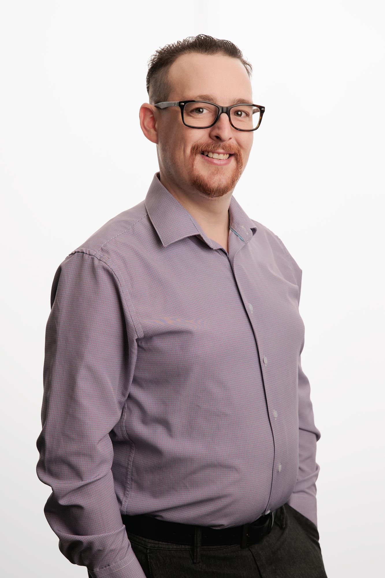 Team headshots Calgary studio: Smiling professional man in a lavender micro-check button-down shirt and dark-frame glasses in a relaxed three-quarter pose, white background