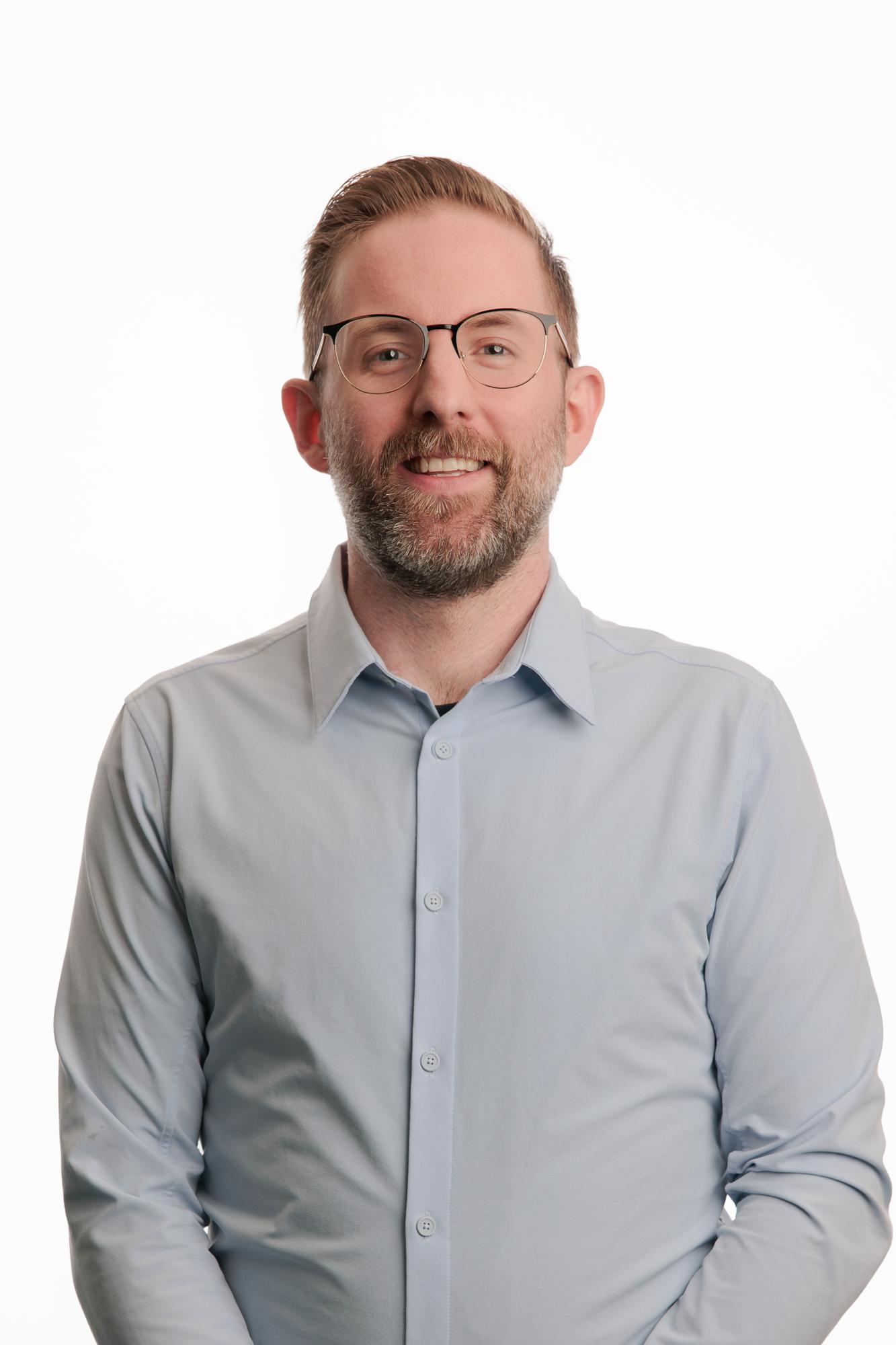 Team headshots Calgary studio: Approachable professional man with a beard and round metal-frame glasses in a light blue button-down shirt with hands in pockets and a broad smile, white background