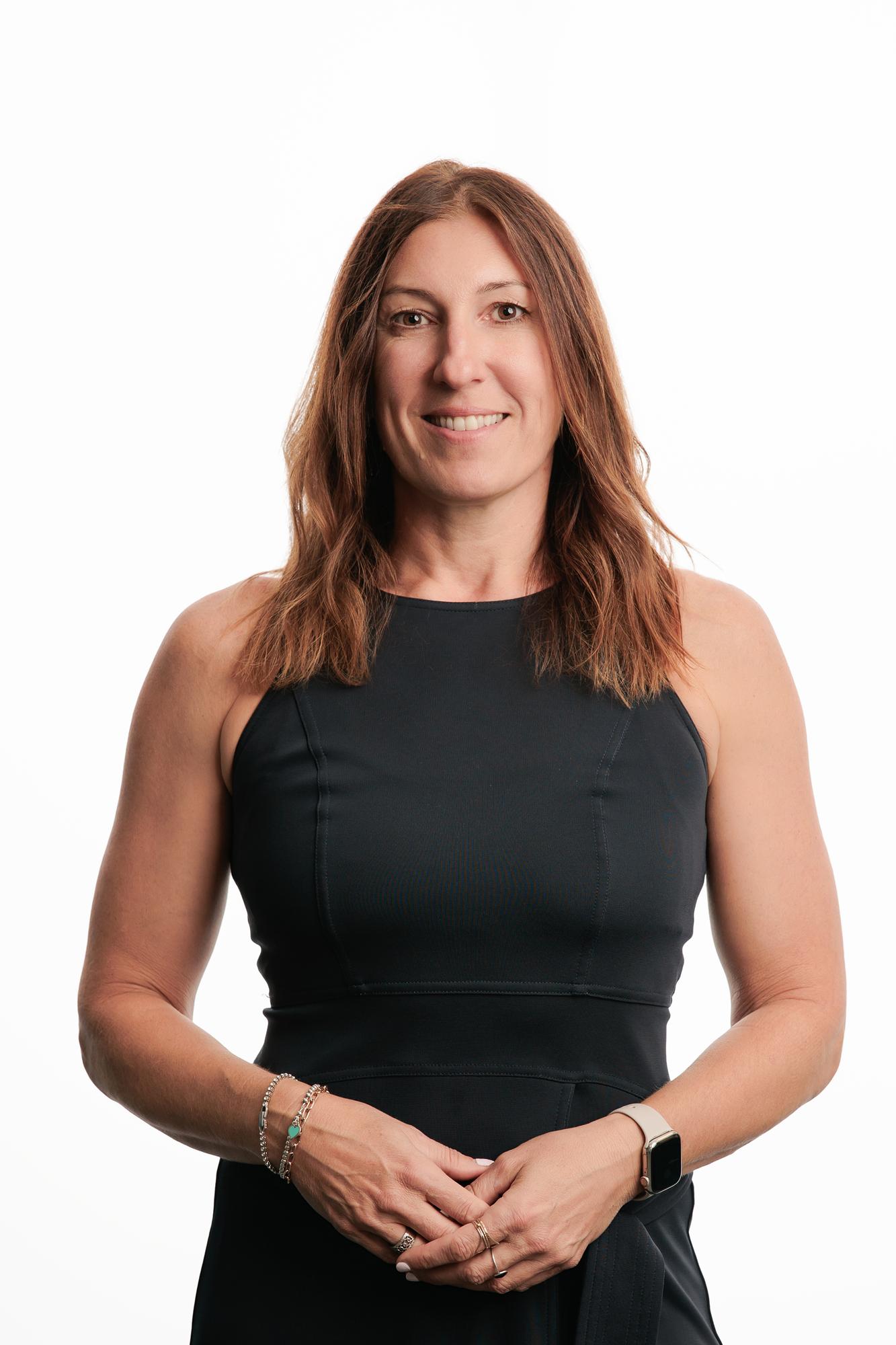 Team headshots Calgary studio: Approachable professional woman with auburn hair in a black sleeveless top and silver bracelet standing confidently against a white studio backdrop