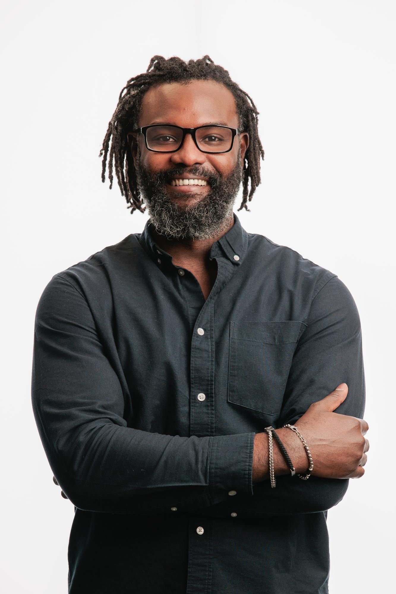 Team headshots Calgary studio: Confident professional man with dreadlocks, a beard, and bold black-frame glasses in a dark teal button-down shirt with arms crossed and a wide smile, silver and cord bracelets visible, white background
