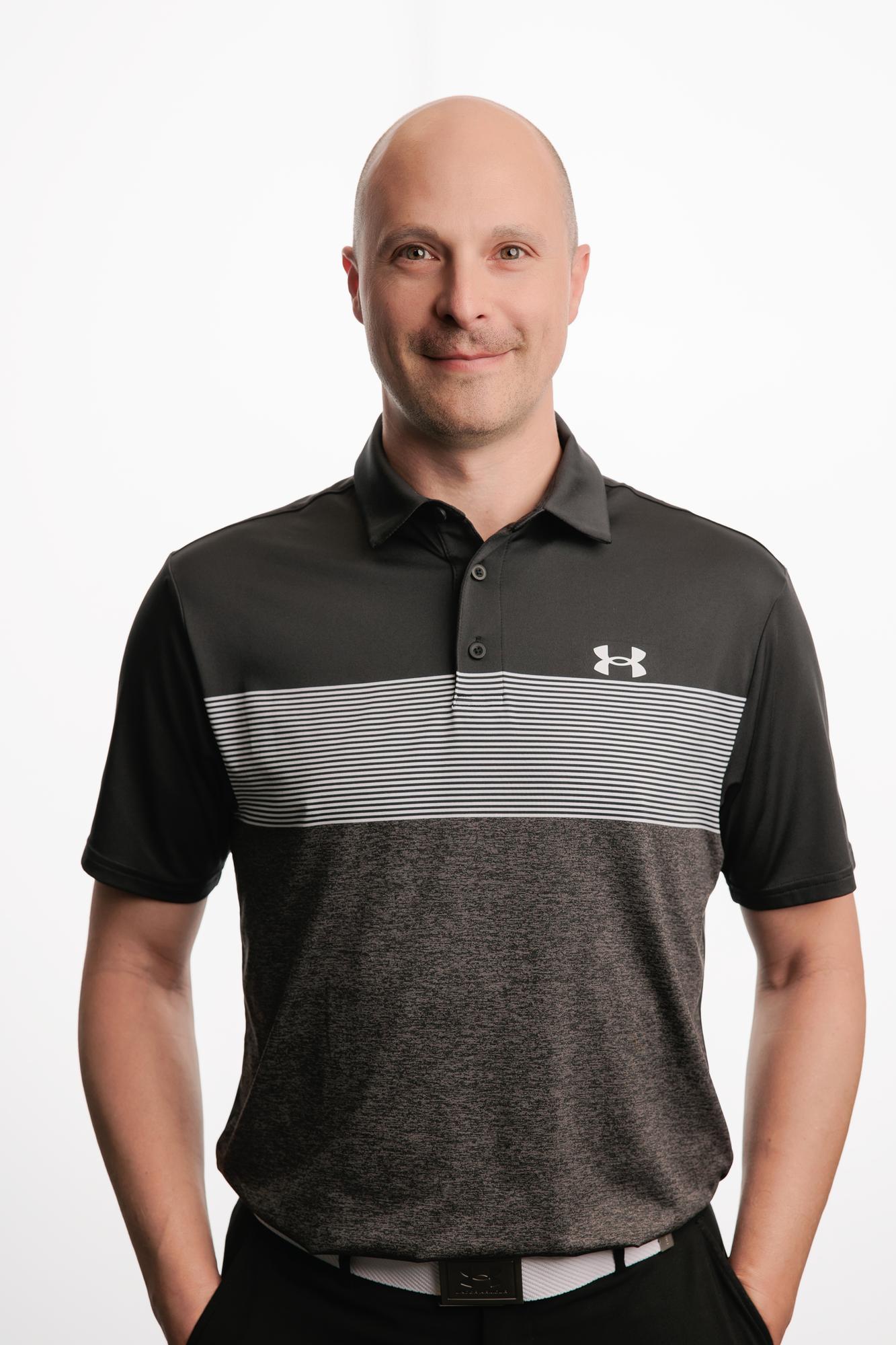 Team headshots Calgary studio: Confident professional man in a black-and-grey striped Under Armour polo with hands in pockets against a white studio backdrop