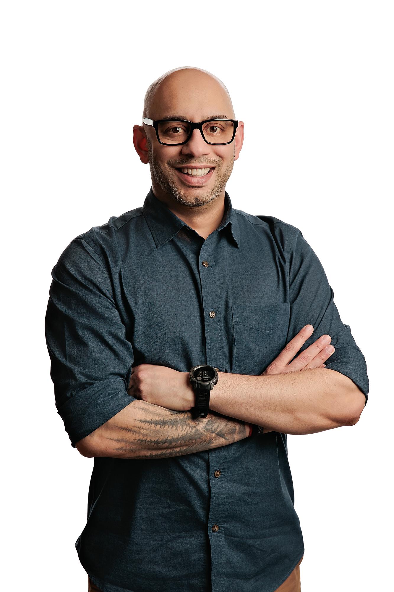 Team headshots Calgary studio: Energetic professional man with a shaved head, goatee, and bold black-frame glasses in a dark teal chambray shirt with arms crossed, a sports watch, and a forearm tattoo, white background