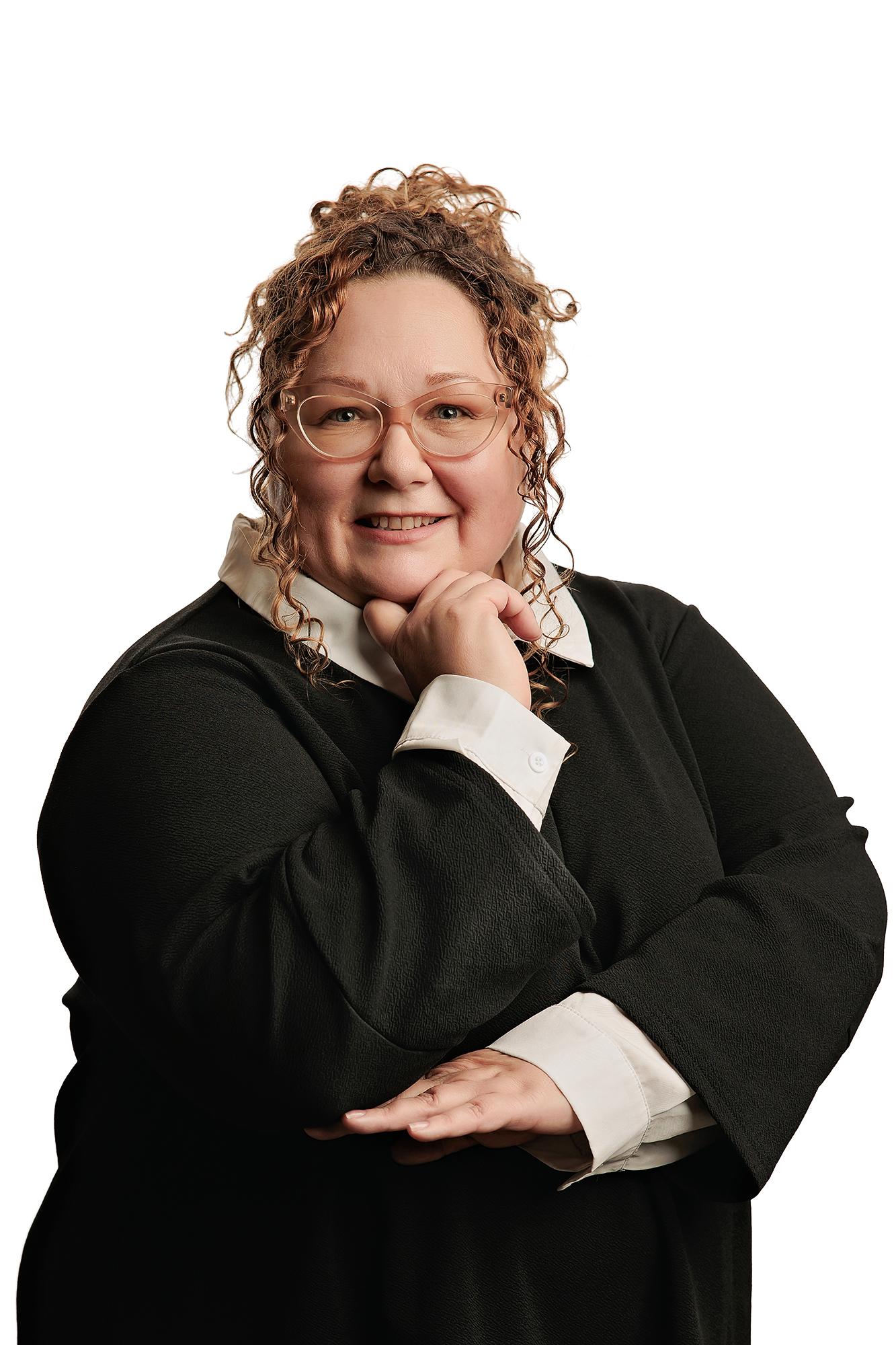Team headshots Calgary studio: Warm and approachable professional woman with auburn curly hair pulled up and peach round-frame glasses in a black top with white collar cuffs, chin resting on hand, white background
