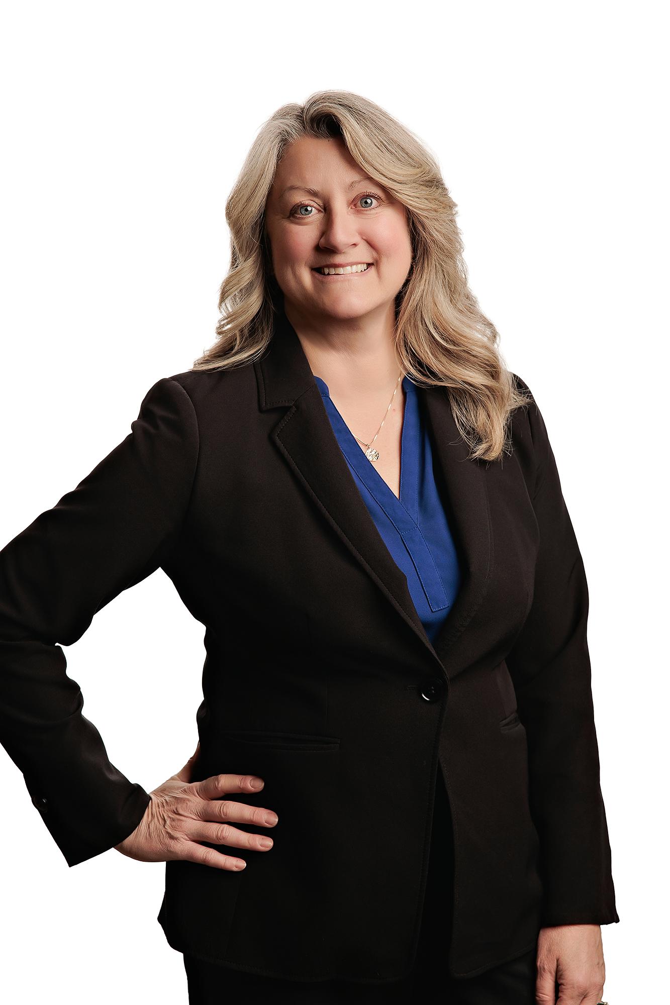 Team headshots Calgary studio: Commanding professional woman with wavy blonde hair in a black blazer over a cobalt blue V-neck blouse with hand on hip and a warm smile, white background