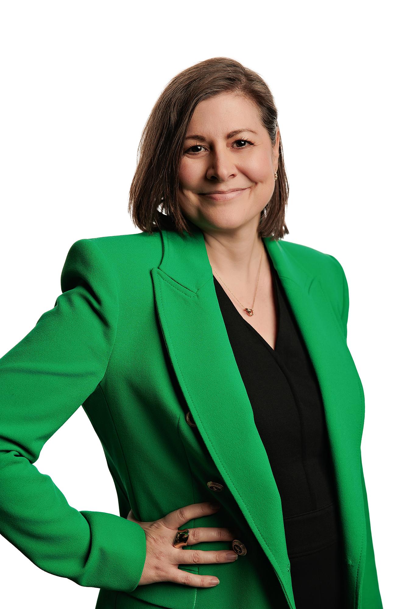 Team headshots Calgary studio: Poised professional woman with a dark bob, gold drop earrings, and a vibrant kelly green blazer over a black V-neck top with hand on hip and a subtle smile, white background