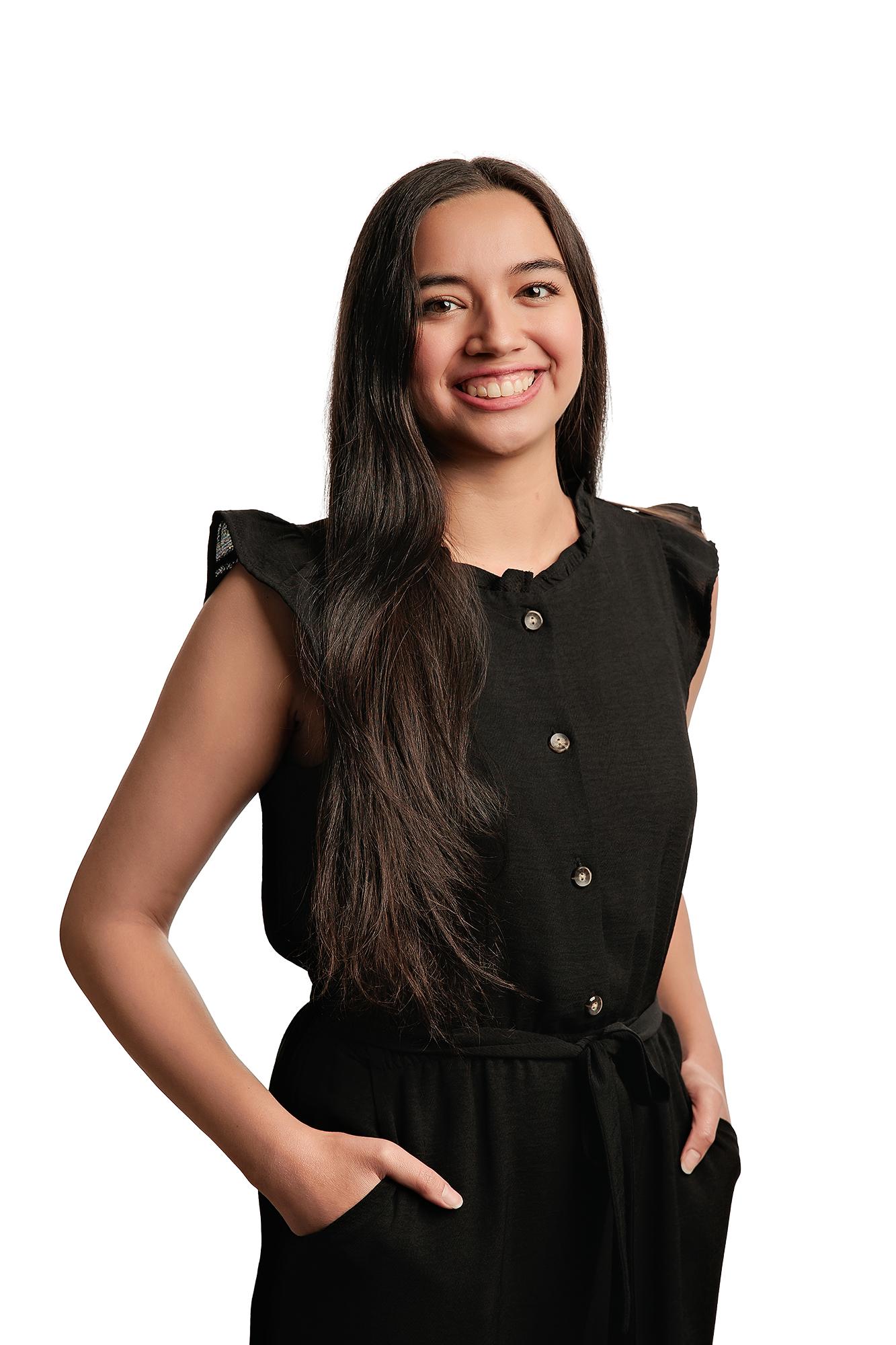 Team headshots Calgary studio: Joyful young professional woman with long dark hair in a black ruffle-sleeve button-front jumpsuit with hands in pockets and a radiant smile, white background