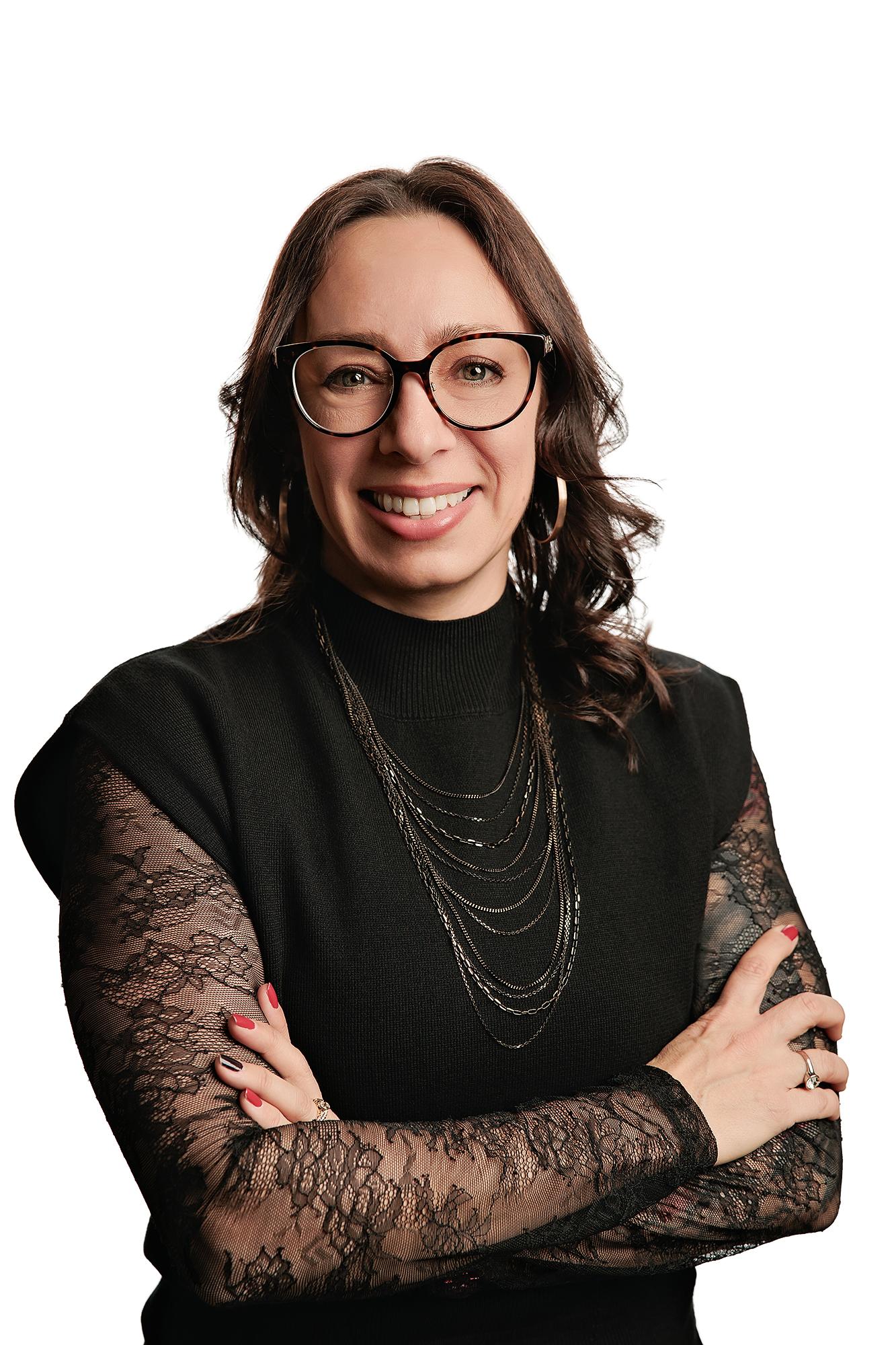 Team headshots Calgary studio: Confident professional woman with dark brown wavy hair and tortoiseshell cat-eye glasses in a black mock-turtleneck top with layered gold chain necklaces and arms crossed, lace sleeves visible, white background