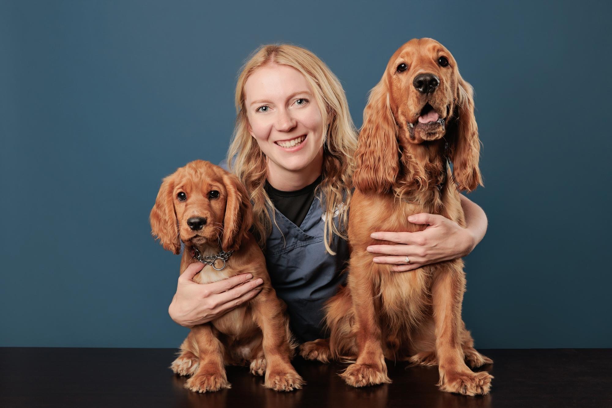 Veterinary team headshots Calgary studio: Smiling female veterinary professional with long blonde wavy hair in blue scrubs posing with two golden Cocker Spaniels — a puppy and an adult — against a deep teal background