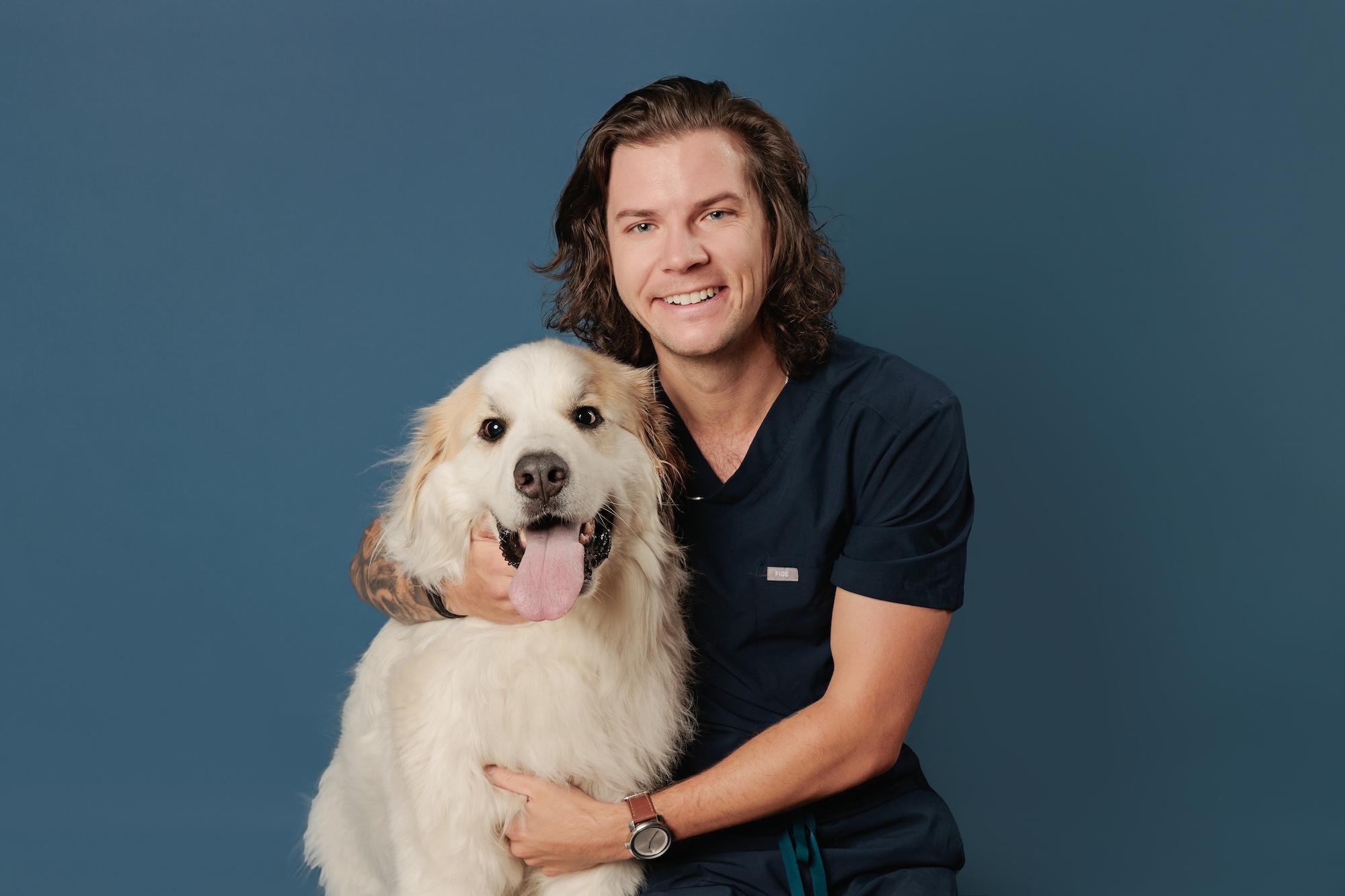 Veterinary team headshots Calgary studio: Smiling male veterinary professional with shoulder-length dark wavy hair in navy scrubs embracing a large fluffy Great Pyrenees dog against a deep teal background, arm tattoo visible