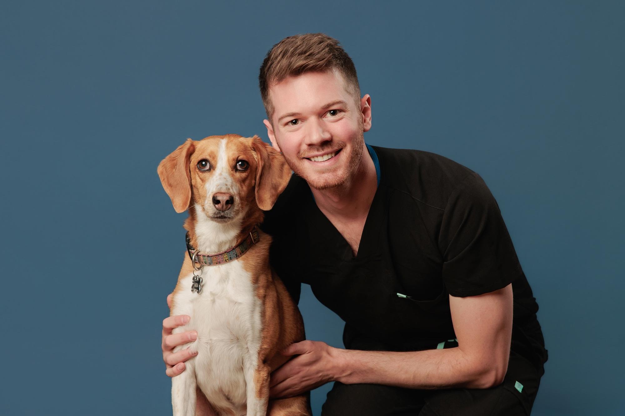 Veterinary team headshots Calgary studio: Smiling male veterinary professional with short auburn hair and a beard in black scrubs crouching beside a medium-sized tan-and-white Beagle mix with a colourful collar against a deep teal background